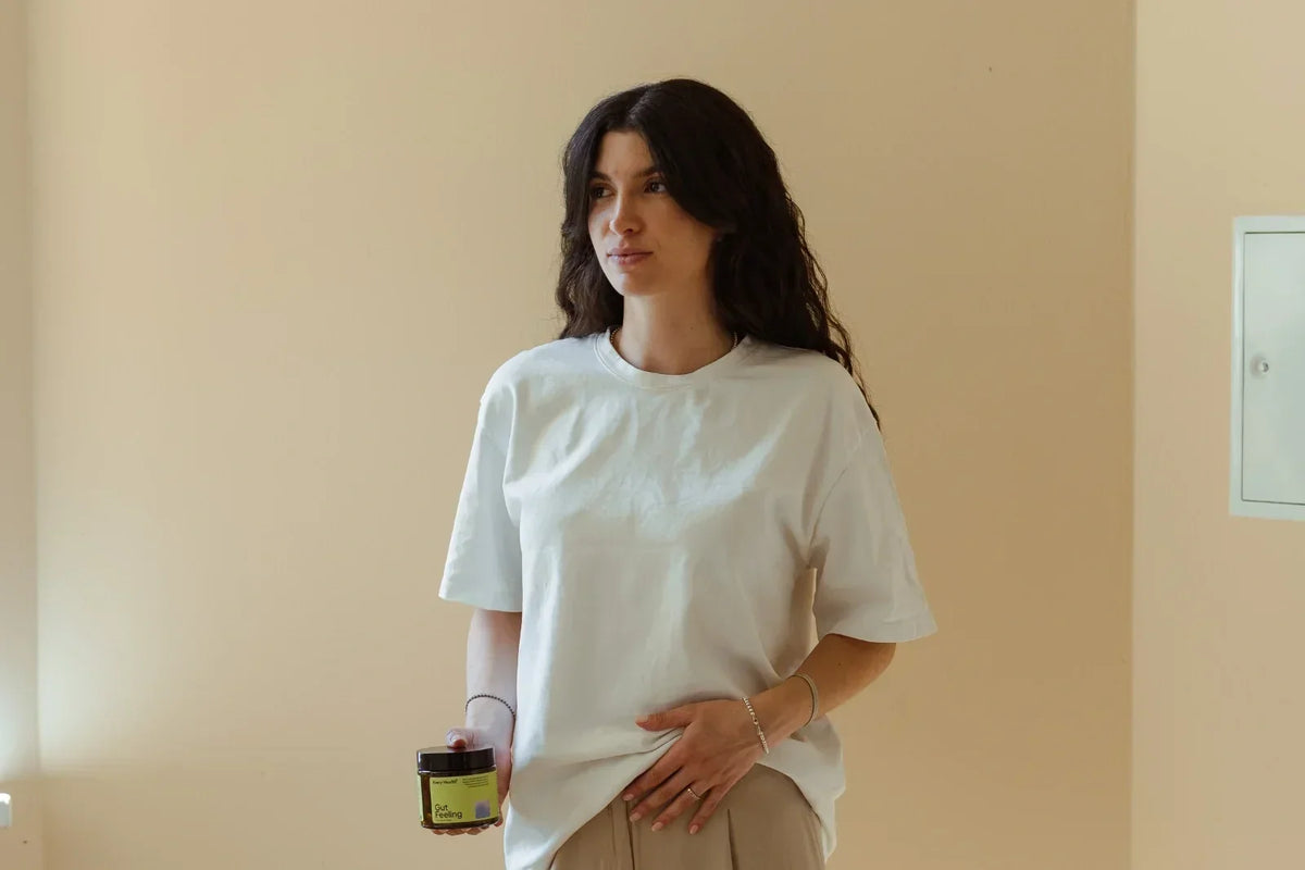 Eine junge Frau in weißem T-Shirt hält ein Gut Feeling Tiegel von Every Health und legt ihre andere Hand sanft auf ihren Bauch.