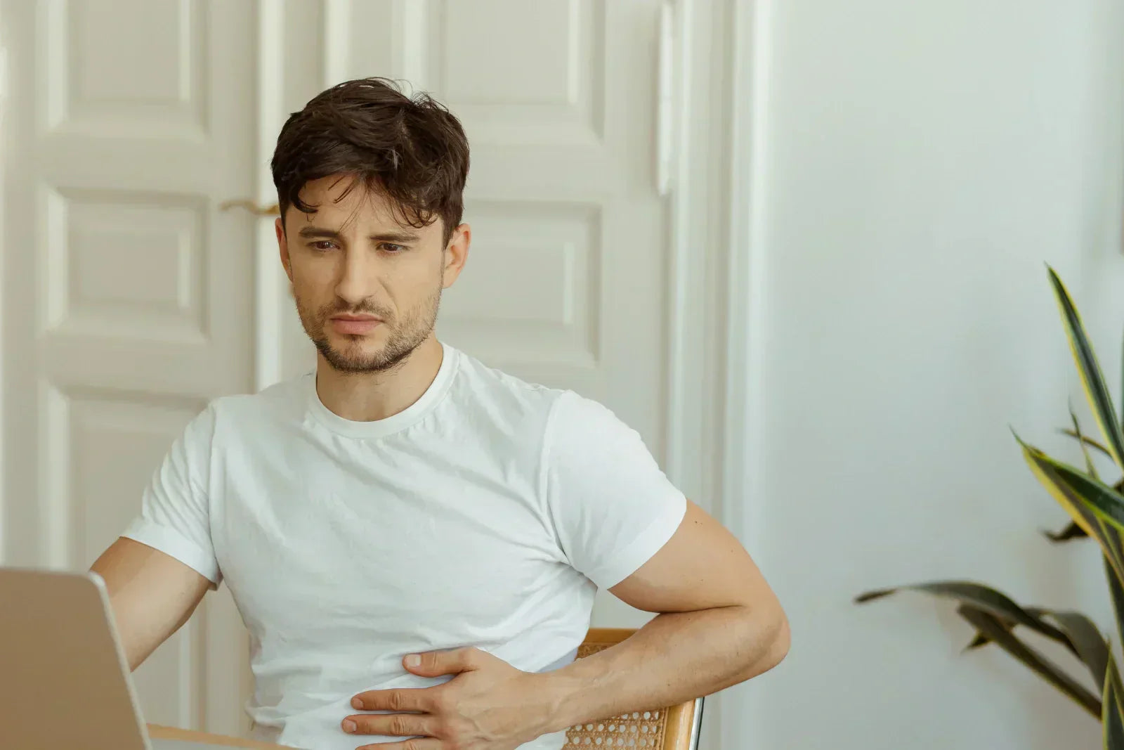 Ein junger Mann sitzt mit schmerzverzerrtem Gesicht am Laptop und hält sich mit der Hand den Bauch.