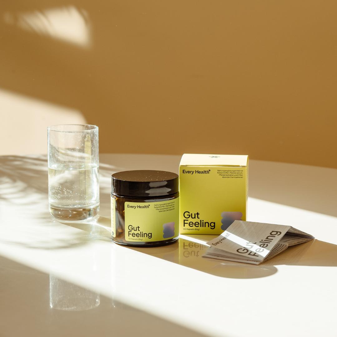 Stillleben des „Gut Feeling“ Produkts von Every Health auf einem weißen, spiegelnden Tisch im warmen Sonnenlicht. Zu sehen sind die braune Kapseldose, die grüne Produktverpackung, eine geöffnete Informationsbroschüre und ein Glas Wasser.