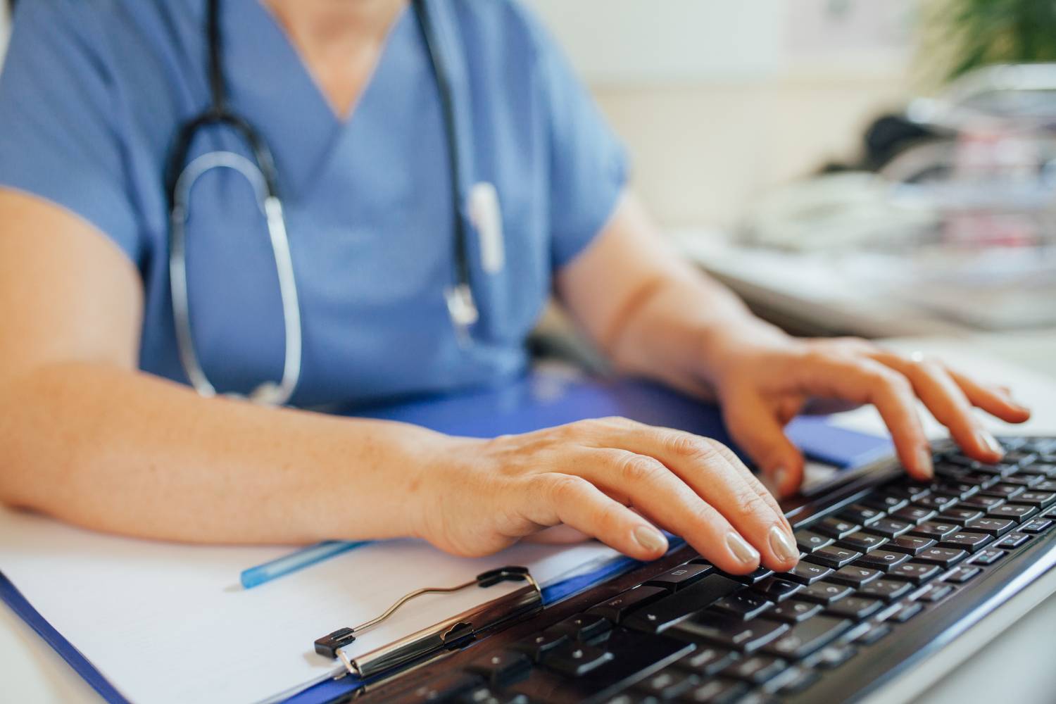 Eine medizinische Fachkraft in blauer Arbeitskleidung mit Stethoskop arbeitet am Computer. Das Bild veranschaulicht die sorgfältige ärztliche Prüfung des Online-Fragebogens zur Behandlung von erektiler Dysfunktion.