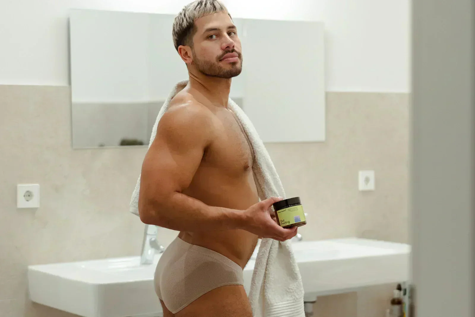 Ein Mann mit kurzen, blondierten Haaren und Bart steht in Unterwäsche im Badezimmer. Er hat ein weißes Handtuch über die Schulter gelegt, blickt in die Kamera und hält eine Produktose mit der Aufschrift „Gut Feeling“ in der Hand.