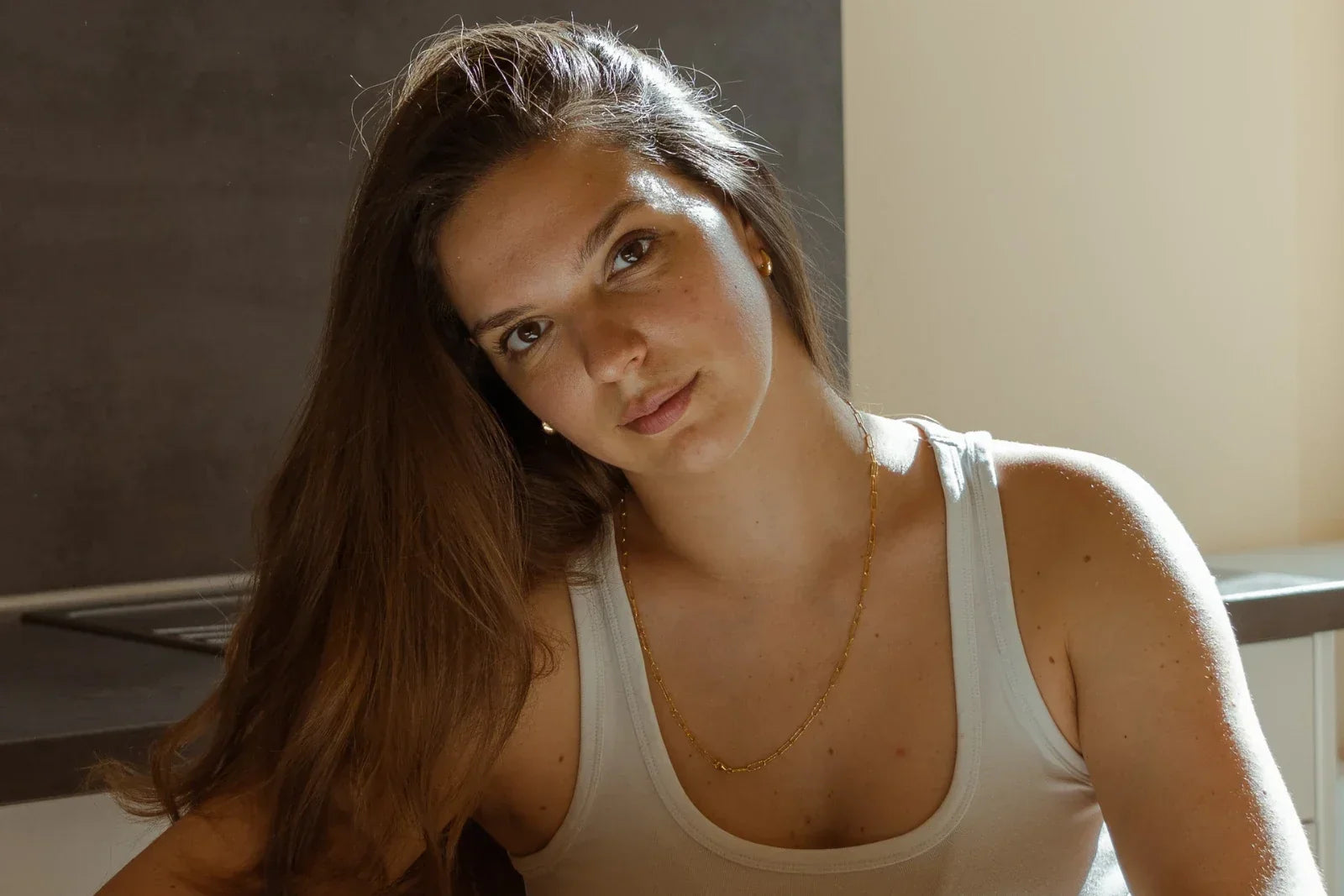 Porträt einer jungen Frau mit langen braunen Haaren und weißem Trägershirt, die an einem Tisch in einer Küche sitzt.