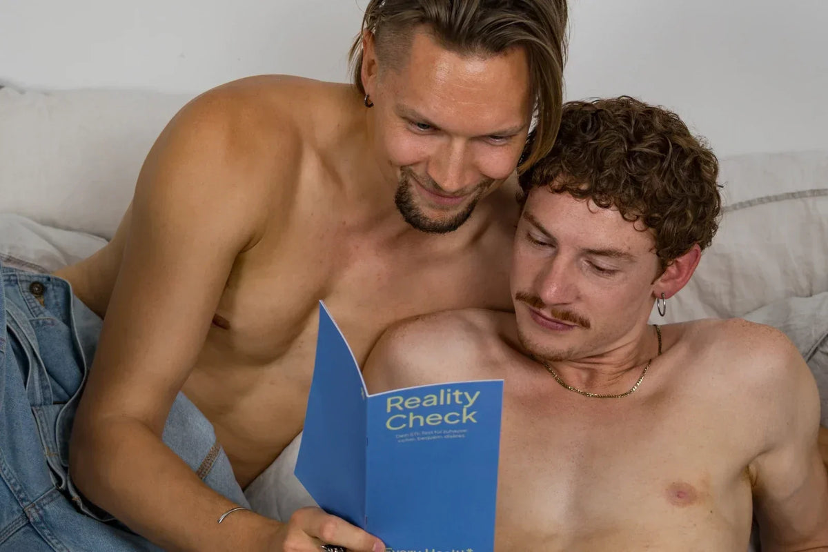 Zwei Männer mit freiem Oberkörper liegen im Bett und lesen gemeinsam eine blaue Broschüre mit der Aufschrift „Reality Check“.