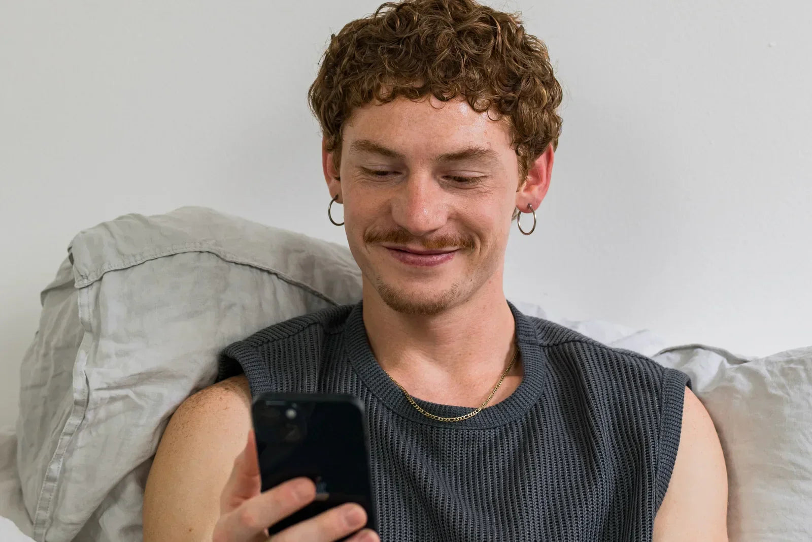 Ein lächelnder Mann mit lockigen, rötlich-braunen Haaren sitzt im Bett und schaut auf sein Smartphone.