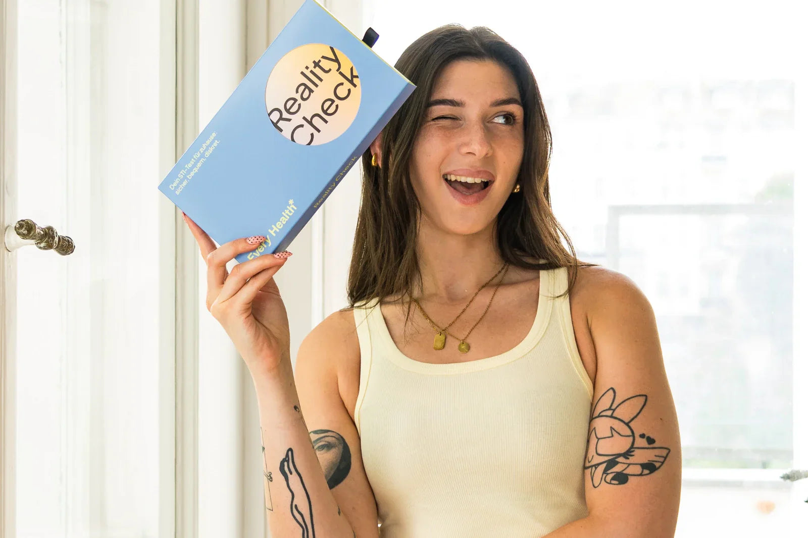 Eine Frau mit Tattoos am Arm zwinkert fröhlich und hält eine hellblaue Schachtel mit der Aufschrift 'Reality Check' hoch.