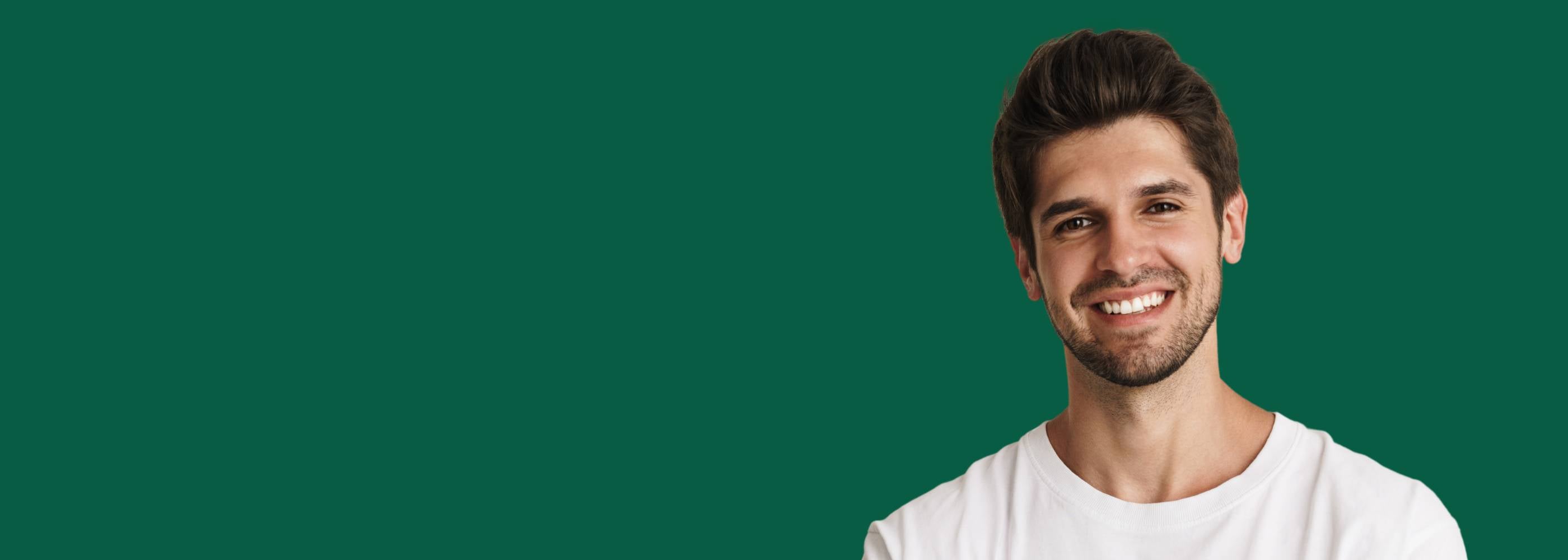 Nahaufnahme eines lächelnden Mannes mit dunklen Haaren und Bart in einem weißen T-Shirt vor einem dunkelgrünen Hintergrund. Er blickt positiv in die Kamera. Das Bild visualisiert die positive Auswirkung und wiedergewonnene Lebensfreude nach der diskreten Behandlung von Erektionsstörungen.