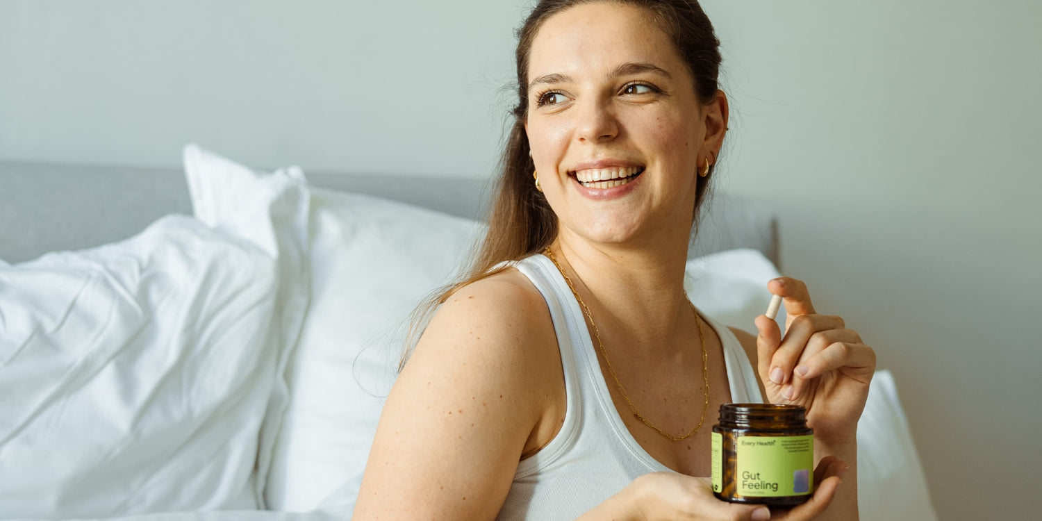 Eine lächelnde junge Frau sitzt im Bett und hält eine geöffnete Dose „Gut Feeling“ von Every Health in der Hand, während sie eine Kapsel zur Einnahme bereithält.