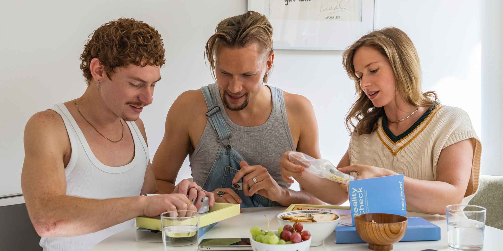 Drei junge Leute sitzen an einem Tisch und packen gemeinsam eine Box mit der Aufschrift „Reality Check“ aus. Auf dem Tisch stehen gesunde Snacks und Wasser; die Stimmung ist fokussiert und freundschaftlich.